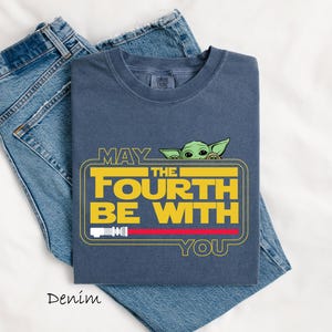 May include: A denim blue t-shirt with the text "MAY THE FOURTH BE WITH YOU" in yellow and white, featuring a green cartoon character. The shirt is folded on top of a pair of blue jeans.