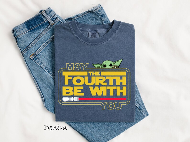 May include: A denim blue t-shirt with the text "MAY THE FOURTH BE WITH YOU" in yellow and white, featuring a green cartoon character. The shirt is folded on top of a pair of blue jeans.