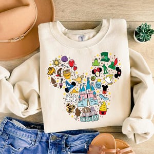 Puede incluir: Sudadera color crema con un dise&ntilde;o de cabeza de Mickey Mouse hecho de varios iconos de dibujos animados. El dise&ntilde;o incluye un castillo, helado y el texto "it's a small world".