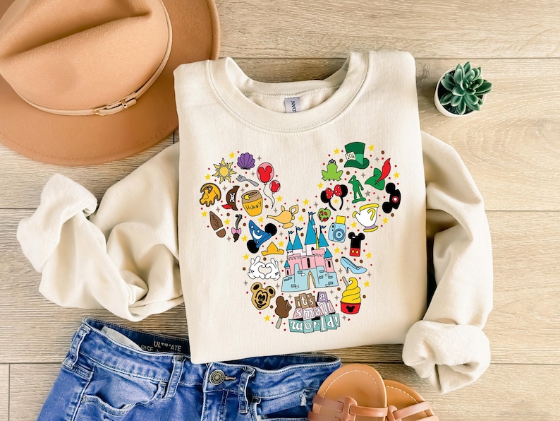 Puede incluir: Sudadera color crema con un dise&ntilde;o de cabeza de Mickey Mouse hecho de varios iconos de dibujos animados. El dise&ntilde;o incluye un castillo, helado y el texto "it's a small world".
