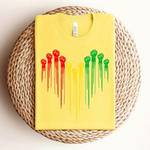 May include: Yellow t-shirt with a crew neck featuring a graphic of raised fists in red, yellow, and green, with dripping paint effect. The shirt is made of a soft material and is folded on a woven surface.