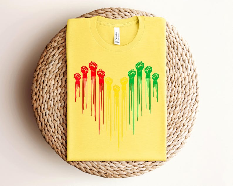 May include: Yellow t-shirt with a crew neck featuring a graphic of raised fists in red, yellow, and green, with dripping paint effect. The shirt is made of a soft material and is folded on a woven surface.