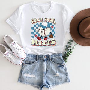 May include: White t-shirt with a retro baseball graphic and the text "Calm Your Mitts". The shirt is paired with denim shorts and white sneakers.
