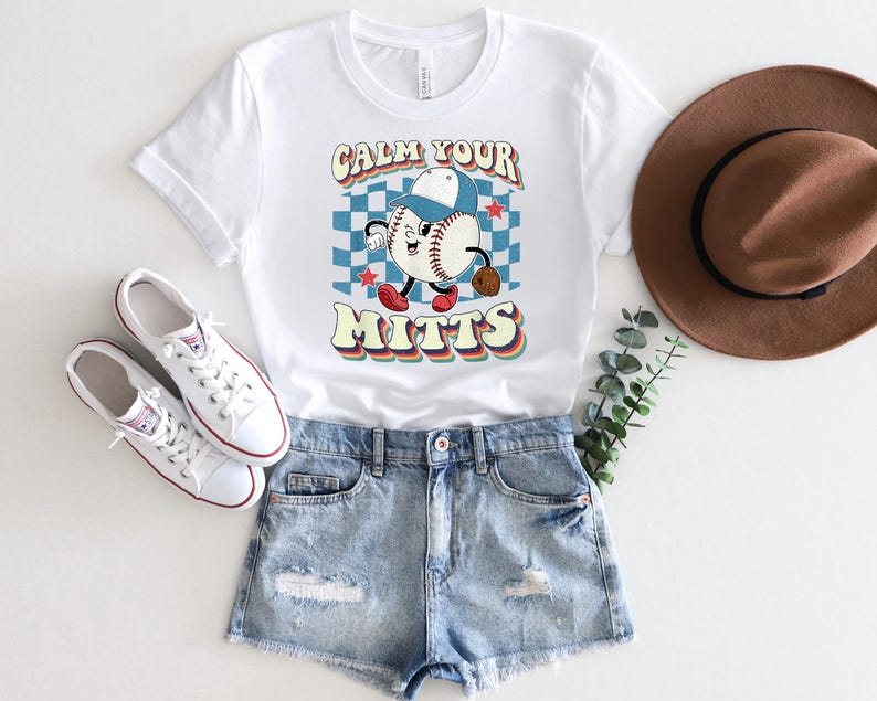 May include: White t-shirt with a retro baseball graphic and the text "Calm Your Mitts". The shirt is paired with denim shorts and white sneakers.