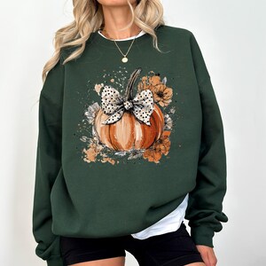 May include: Dark green sweatshirt featuring a watercolor-style pumpkin with a polka-dot bow and floral accents. The pumpkin is orange with a green stem. The design is centered on the front of the long-sleeved top.
