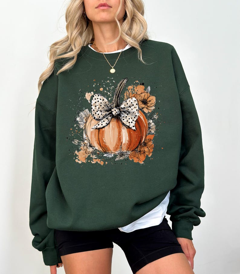 May include: Dark green sweatshirt featuring a watercolor-style pumpkin with a polka-dot bow and floral accents. The pumpkin is orange with a green stem. The design is centered on the front of the long-sleeved top.