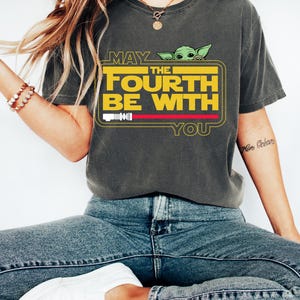 May include: Dark gray t-shirt with the text "MAY THE FOURTH BE WITH YOU" in yellow and white, featuring a green cartoon character. The shirt has a red stripe and a white bar graphic. The shirt is worn with blue jeans and white shoes.