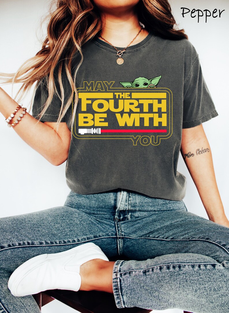 May include: Dark gray t-shirt with the text "MAY THE FOURTH BE WITH YOU" in yellow and white, featuring a green cartoon character. The shirt has a red stripe and a white bar graphic. The shirt is worn with blue jeans and white shoes.