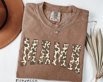 May include: A brown t-shirt with a camo print "MAMA" graphic. The shirt is a relaxed fit and has a distressed look. The shirt is shown on a flat surface with a brown cowboy hat and a sprig of dried grass.