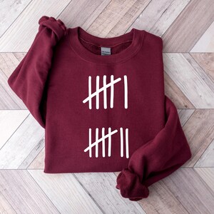 May include: A maroon sweatshirt with white tally marks printed on the front. The design consists of two sets of vertical lines, each with a single diagonal line through them. The sweatshirt is folded on a light wood-grain surface.