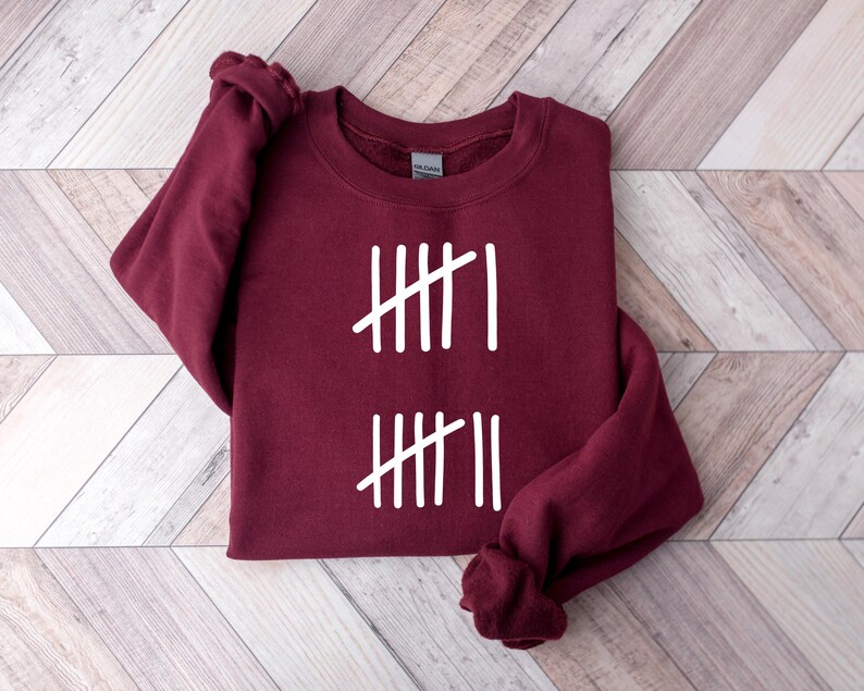 May include: A maroon sweatshirt with white tally marks printed on the front. The design consists of two sets of vertical lines, each with a single diagonal line through them. The sweatshirt is folded on a light wood-grain surface.