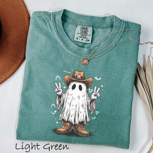 Pu&ograve; includere: T-shirt verde chiaro con una grafica di un fantasma con cappello e stivali da cowboy, che fa il segno della pace. La maglietta ha un girocollo e maniche corte. Il testo "Light Green" &egrave; in basso nell'immagine.