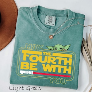May include: Light green t-shirt with the text "MAY THE FOURTH BE WITH YOU" in yellow, featuring a green cartoon character. The shirt has a crew neck and short sleeves. A silver necklace with a pendant is draped across the shirt.