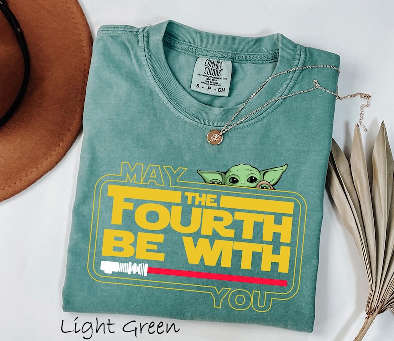 May include: Light green t-shirt with the text "MAY THE FOURTH BE WITH YOU" in yellow, featuring a green cartoon character. The shirt has a crew neck and short sleeves. A silver necklace with a pendant is draped across the shirt.