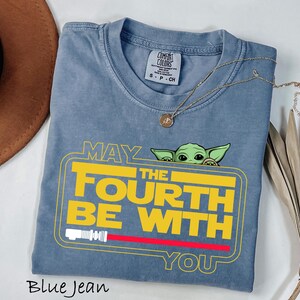 May include: Blue jean colored t-shirt with the text "MAY THE FOURTH BE WITH YOU" in yellow and white, with a red lightsaber graphic. A small green alien character peeks from behind the text. A gold necklace rests on the shirt.