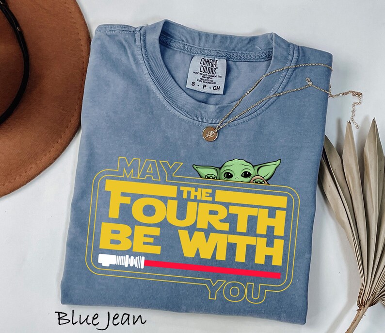 May include: Blue jean colored t-shirt with the text "MAY THE FOURTH BE WITH YOU" in yellow and white, with a red lightsaber graphic. A small green alien character peeks from behind the text. A gold necklace rests on the shirt.