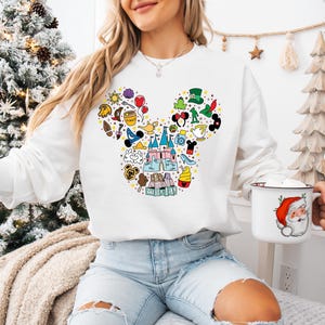 Puede incluir: Sudadera blanca con un dise&ntilde;o colorido de la cabeza de Mickey Mouse llena de varios iconos de Disney. La sudadera se combina con vaqueros azul claro. Tambi&eacute;n se ve una taza blanca con un gr&aacute;fico de Pap&aacute; Noel.