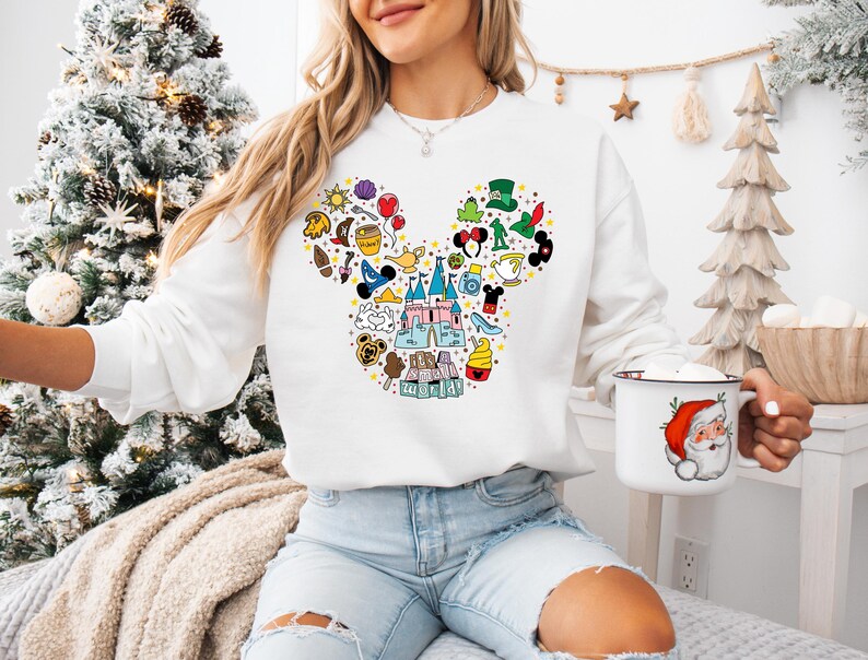 Puede incluir: Sudadera blanca con un dise&ntilde;o colorido de la cabeza de Mickey Mouse llena de varios iconos de Disney. La sudadera se combina con vaqueros azul claro. Tambi&eacute;n se ve una taza blanca con un gr&aacute;fico de Pap&aacute; Noel.