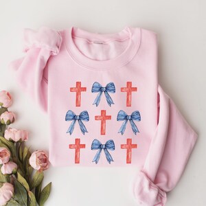 May include: A pink sweatshirt featuring a design of red crosses and blue bows. The design is repeated across the front of the sweatshirt.