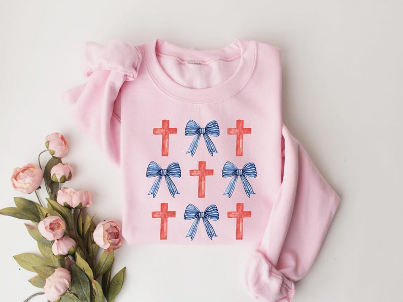 May include: A pink sweatshirt featuring a design of red crosses and blue bows. The design is repeated across the front of the sweatshirt.