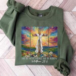 May include: Green sweatshirt featuring a colourful painting of a cross with a white cloth draped over it. The painting has a sunset backdrop with green hills and flowers. The text "He is not here. He is risen. Matthew 28:6" is printed below the painting.