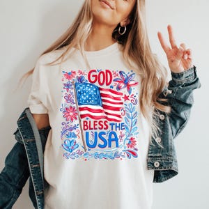 May include: A cream-colored t-shirt featuring a graphic print of an American flag on a pole with the text GOD BLESS THE USA in red and blue. The design is surrounded by decorative red and blue floral and leafy patterns on a white background.