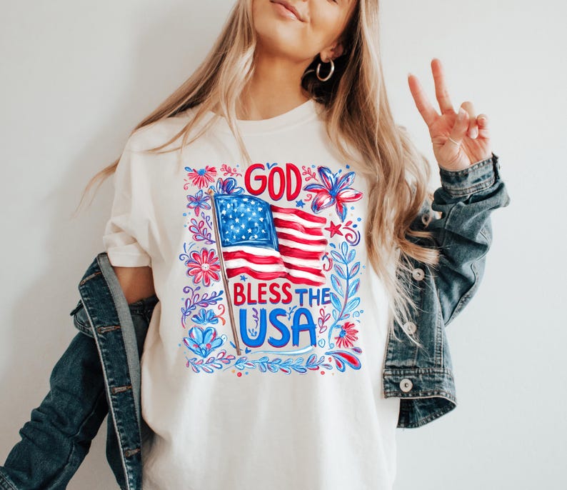 May include: A cream-colored t-shirt featuring a graphic print of an American flag on a pole with the text GOD BLESS THE USA in red and blue. The design is surrounded by decorative red and blue floral and leafy patterns on a white background.