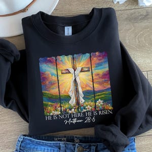 May include: Black sweatshirt featuring a colourful painting of a cross and the text "HE IS NOT HERE. HE IS RISEN. Matthew 28:6".