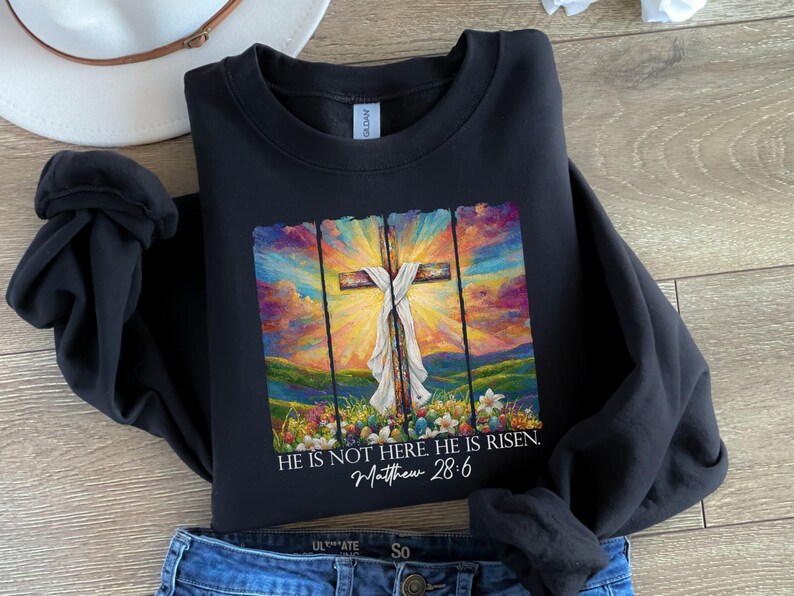 May include: Black sweatshirt featuring a colourful painting of a cross and the text "HE IS NOT HERE. HE IS RISEN. Matthew 28:6".