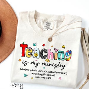 May include: Ivory-colored t-shirt with the words "Teaching is my ministry" in colorful, playful lettering. The shirt includes the Bible verse Colossians 3:23. A gold necklace and a brown hat are also visible.