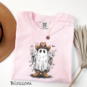 Pu&ograve; includere: T-shirt rosa chiaro con una grafica di un fantasma con cappello e stivali da cowboy, che fa il segno della pace. La parola "Blossom" &egrave; in basso. Una collana d'oro con un ciondolo &egrave; sulla maglietta.