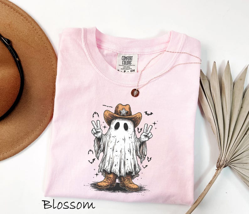 Pu&ograve; includere: T-shirt rosa chiaro con una grafica di un fantasma con cappello e stivali da cowboy, che fa il segno della pace. La parola "Blossom" &egrave; in basso. Una collana d'oro con un ciondolo &egrave; sulla maglietta.