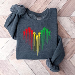 May include: A dark gray sweatshirt featuring a heart-shaped design of raised fists in red, yellow, and green. The fists have a dripping effect, creating a unique visual. The sweatshirt is made of a soft material.