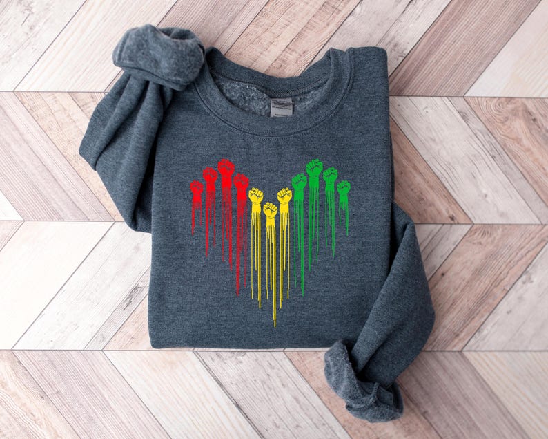May include: A dark gray sweatshirt featuring a heart-shaped design of raised fists in red, yellow, and green. The fists have a dripping effect, creating a unique visual. The sweatshirt is made of a soft material.