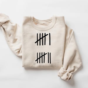 May include: Cream-colored sweatshirt with black tally marks. The design features two sets of vertical lines, with one set having six marks and the other set having seven marks. The sleeves are rolled up, and the sweatshirt is laid flat.