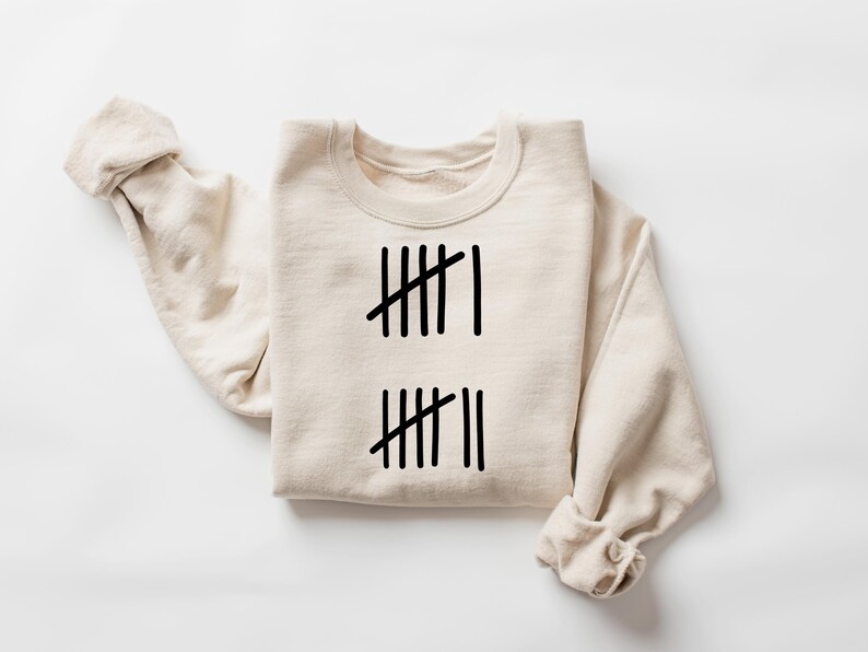 May include: Cream-colored sweatshirt with black tally marks. The design features two sets of vertical lines, with one set having six marks and the other set having seven marks. The sleeves are rolled up, and the sweatshirt is laid flat.