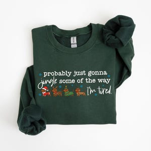 May include: A dark green sweatshirt with the text "probably just gonna jingle some of the way I'm tired." The graphic includes Santa, reindeer, a Christmas tree, and a dachshund. The sweatshirt has long sleeves.