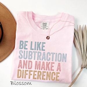 May include: A light pink t-shirt with the text "BE LIKE SUBTRACTION AND MAKE A DIFFERENCE" in blue, pink, and tan. A gold necklace with a small pendant rests on the shirt. The word "Blossom" is printed at the bottom.