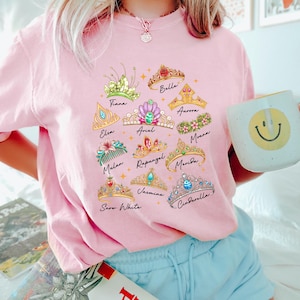 May include: A light pink t-shirt featuring illustrations of various princess tiaras with names like Elsa, Ariel, and Cinderella. The tiaras are in different colors and designs. The shirt is worn by a person.