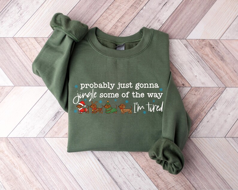 May include: An olive green sweatshirt with the text "probably just gonna jingle some of the way I'm tired." The graphic features dachshunds dressed in Christmas attire. The sweatshirt is folded on a light wood-grain surface.