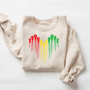 May include: Beige crewneck sweatshirt featuring a heart-shaped design of raised fists in red, yellow, and green. The fists have a dripping paint effect. The sweatshirt is folded with the sleeves rolled up.