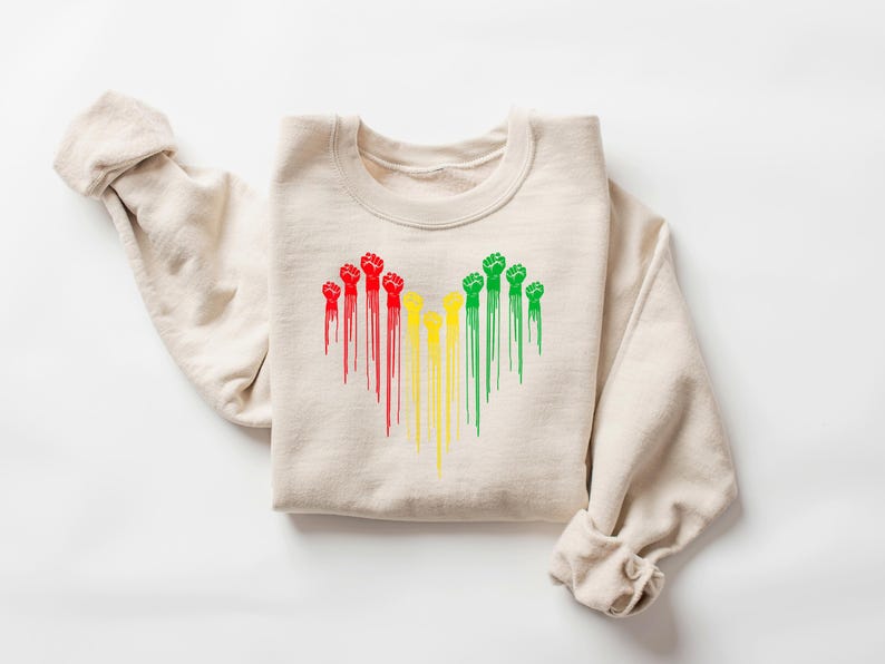 May include: Beige crewneck sweatshirt featuring a heart-shaped design of raised fists in red, yellow, and green. The fists have a dripping paint effect. The sweatshirt is folded with the sleeves rolled up.