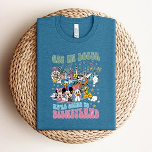 May include: A teal blue t-shirt with a graphic featuring Disney characters and the text "GET IN LOSER WE'RE GOING TO DISNEYLAND" in retro font. The design includes Mickey and Minnie Mouse, Donald Duck, and Goofy.