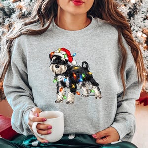 May include: A grey sweatshirt featuring a black and white miniature schnauzer wearing a Santa hat and tangled in Christmas lights.
