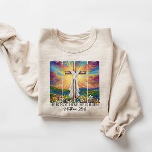 May include: A beige sweatshirt featuring a colourful painting of a cross with a white cloth draped over it. The painting is framed with a wooden border. The text "He is not here He is risen. Matthew 28:6" is printed below the painting.