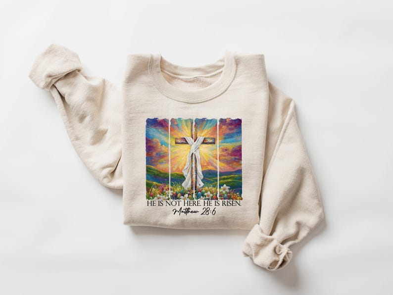 May include: A beige sweatshirt featuring a colourful painting of a cross with a white cloth draped over it. The painting is framed with a wooden border. The text "He is not here He is risen. Matthew 28:6" is printed below the painting.