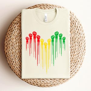 May include: Cream-colored t-shirt featuring a graphic of raised fists in red, yellow, and green, with dripping paint effect. The shirt is displayed on a woven, circular surface.