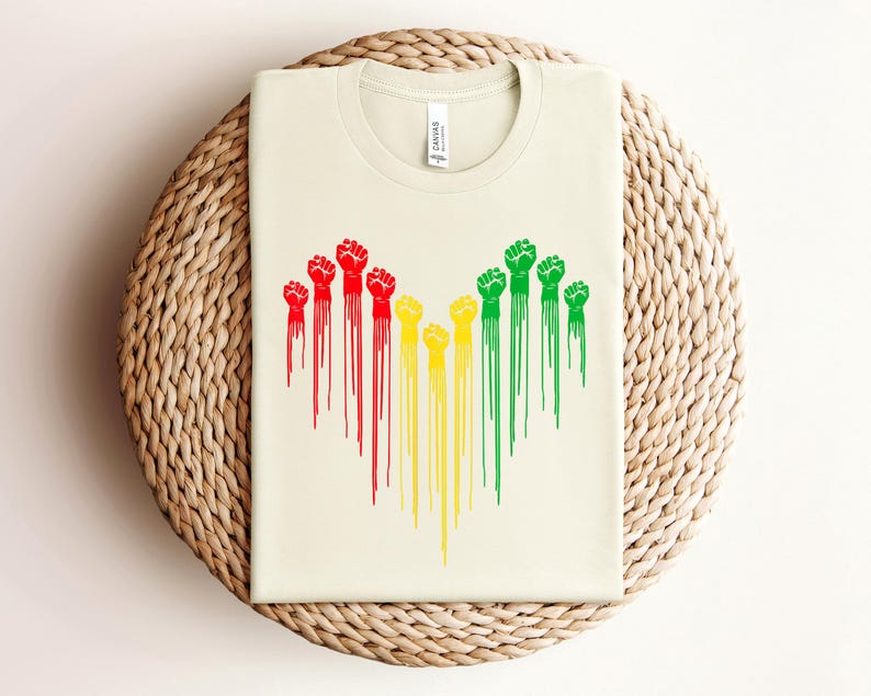 May include: Cream-colored t-shirt featuring a graphic of raised fists in red, yellow, and green, with dripping paint effect. The shirt is displayed on a woven, circular surface.