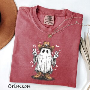 Pu&ograve; includere: T-shirt color cremisi con un fantasma dei cartoni animati che indossa un cappello e stivali da cowboy, che fa il segno della pace. La grafica &egrave; al centro della maglietta. Una collana d'oro &egrave; drappeggiata sulla maglietta. La maglietta &egrave; realizzata in materiale morbido.