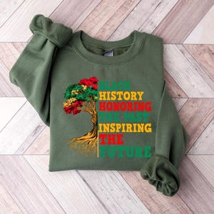 Pu&ograve; includere: Una felpa verde oliva con la scritta "BLACK HISTORY HONORING THE PAST INSPIRING THE FUTURE" in verde, rosso, giallo e verde. Una grafica ad albero con foglie rosse, gialle e verdi &egrave; a sinistra.